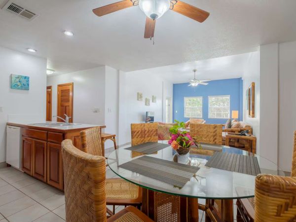 A bright home dining/living area with a glass table, wicker chairs, ceiling fans, and a kitchen counter with wooden cabinets.