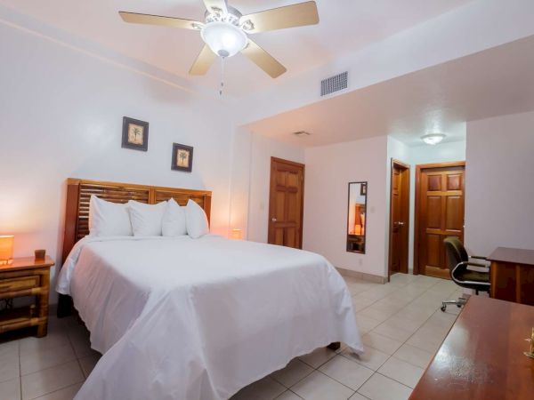 A bright hotel bedroom with a ceiling fan, made bed, bedside lamp, dresser, and a small desk in the corner.