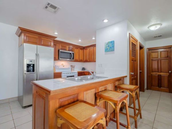 Bright kitchen with wooden cabinets, white fridge, microwave/oven, sink island, and two wooden stools beside hallway doors.
