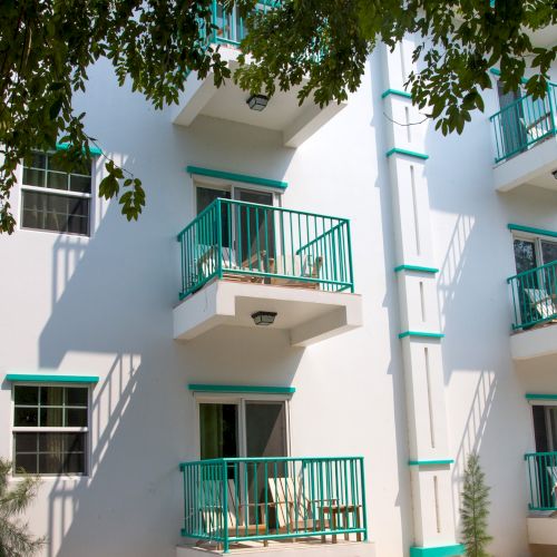 A white apartment building with turquoise railings and balconies, framed by leafy trees overhead.