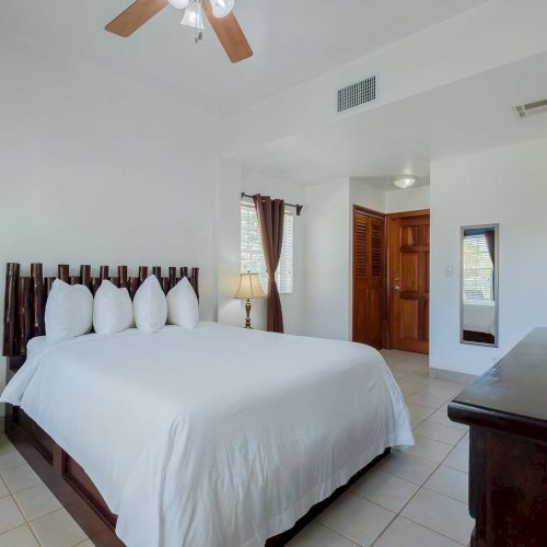 A bright hotel-style bedroom with a king bed, white linens, wooden headboard, bedside lamp, dresser, and tiled floor.