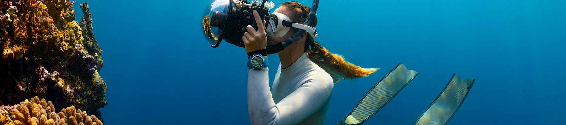 Scuba diver swims above a coral reef, exhaling bubbles in clear blue water.