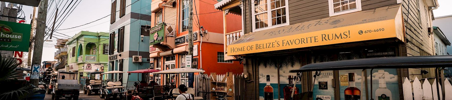 A lively cobblestone street with colorful buildings, parked bikes and carts, and a storefront sign reading &ldquo;Home of Belize&rsquo;s Favorite Rums!&rdquo;.