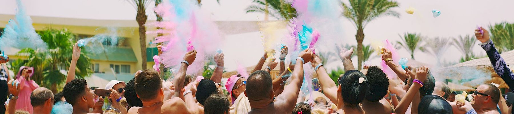 A packed pool party at a sunny beach resort showers the crowd with colorful powder and confetti as everyone cheers.