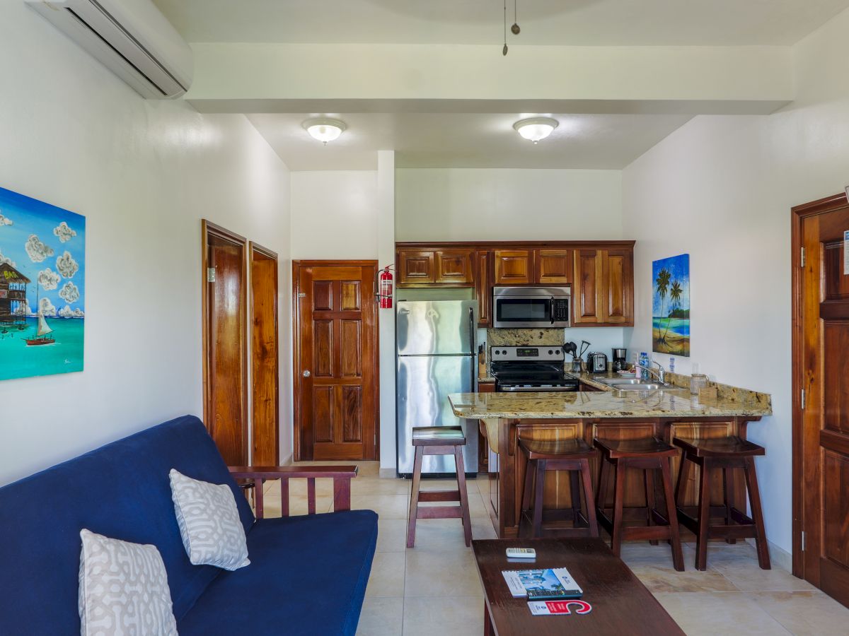 Small lounge kitchen with wooden cabinets, fridge and stove, bar stools, wall art, and an EXIT sign above the door.